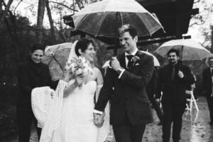 Bride and groom walking together under umbrella during rainy outdoor wedding